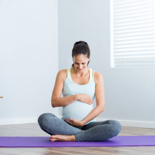 Pregnant woman sitting in lotus position on exercise mat and touching her belly. Pregnant athletic woman ready for yoga prenatal exercise. Hispanic expectant mature mother relaxing at home.